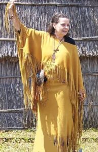 Cheryl Doughty, a woman from Pocomoke Indian Nation. She wears a yellow dress with fringe and has dark brown hair. It looks as if she's in the middle of a dance.