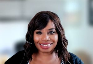 Charmaine Weston, a Black middle-aged woman, with wavy hair past her shoulders that she wears down. She smiles. She wears a black cardigan over a black and white patterned top.